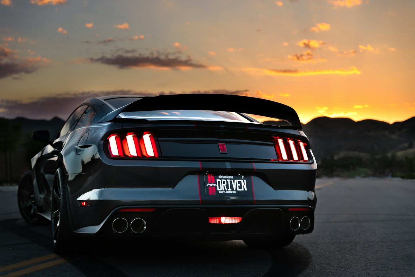 Coche deportivo gris oscuro aparcado en la carretera con luces traseras rojas al atardecer.